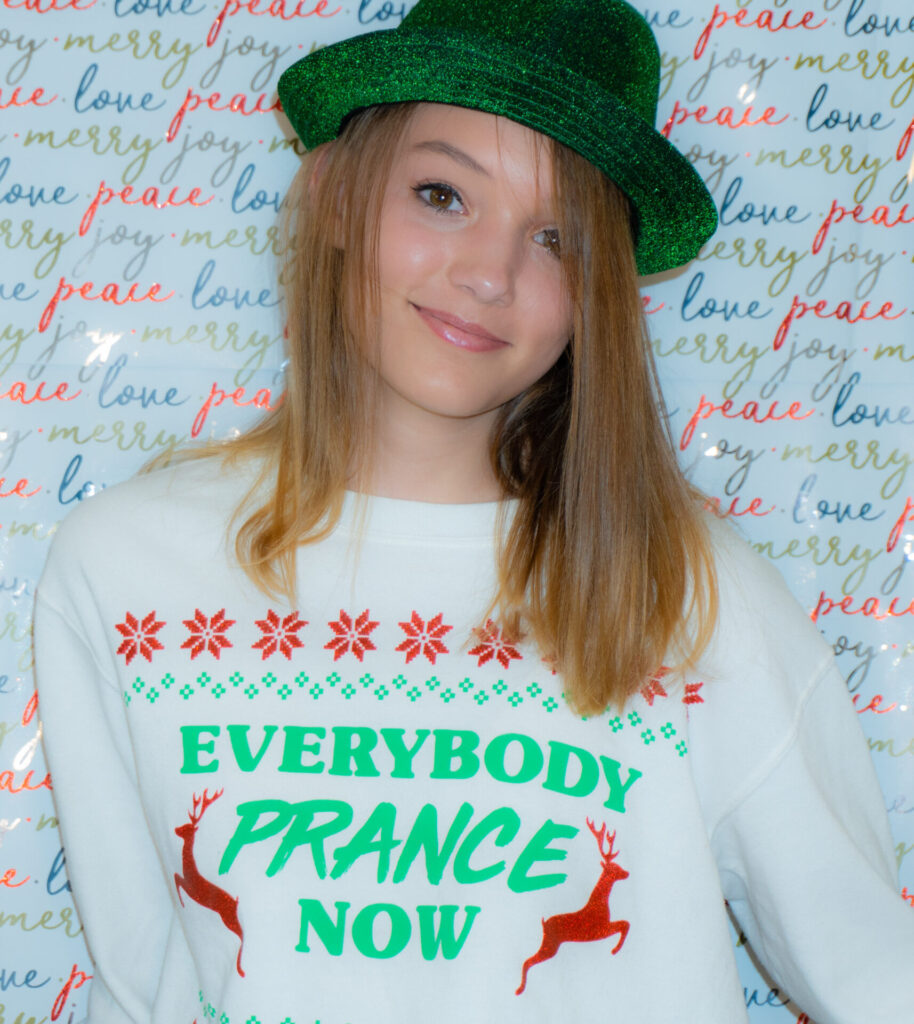 A blonder teen wearing a green fedora, white sweatshirt with christmas colors. Sweatshirt says Everybody Prance now 